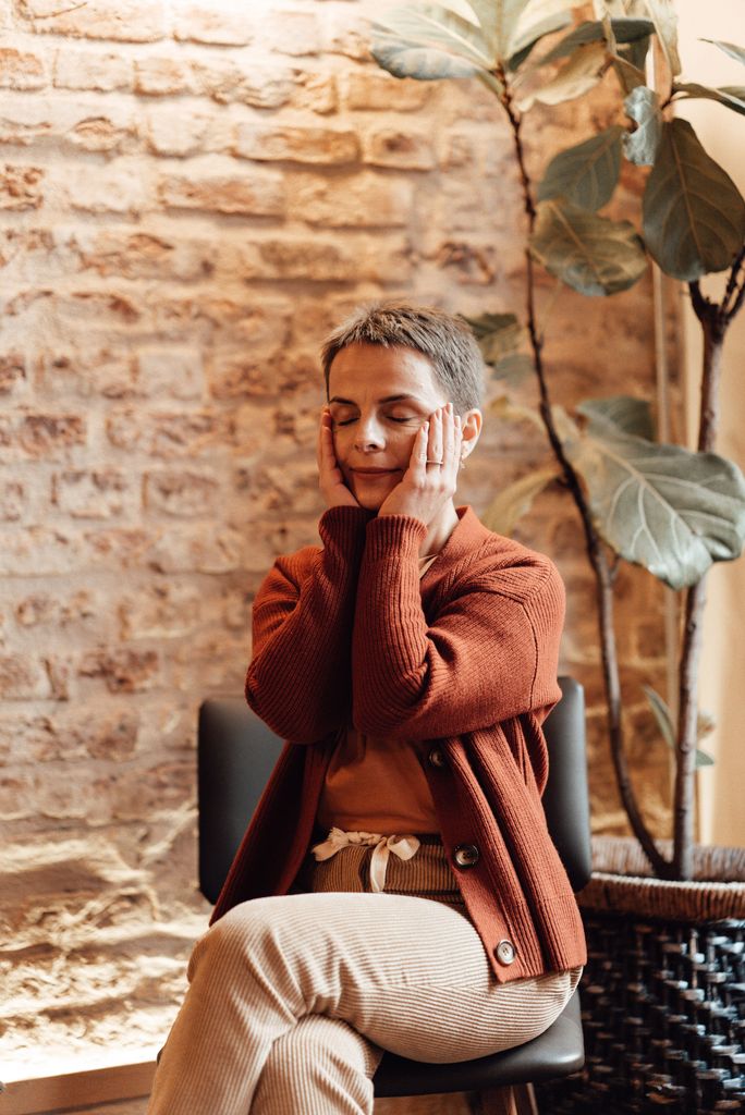 Woman sitting indoors with eyes closed, meditating by a brick wall with a plant, exuding a calming atmosphere.